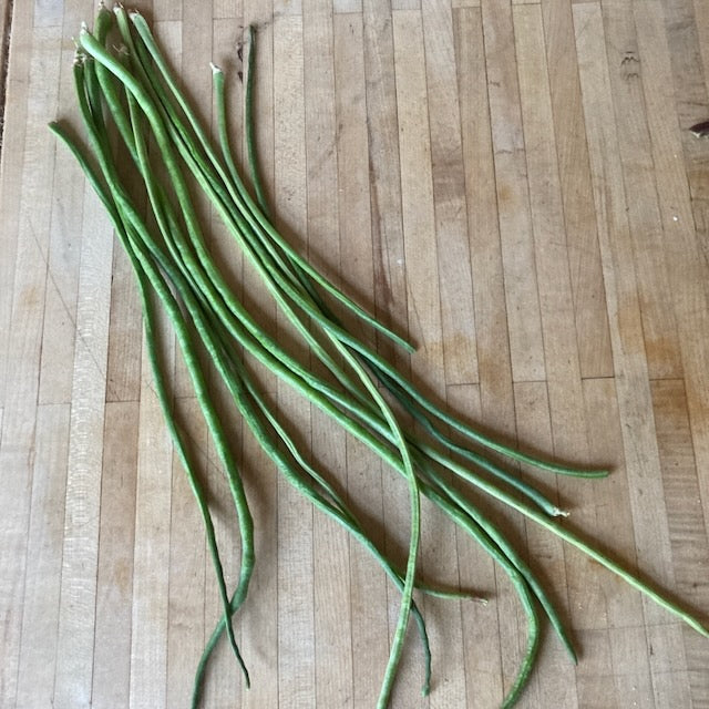 Les fèves Dolique Asperge Liana de Tourne-Sol sont étalées sur un plateau en bois, leur forme élancée, légèrement courbée, contrastant avec les tons chauds du bois.