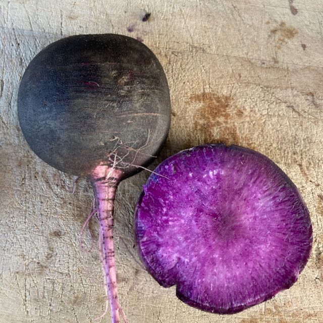 Un Radis d'hiver Améthyste Noir - semences bio de Tourne-Sol est posé à côté d'une coupe transversale de lui-même, révélant un intérieur violet vibrant. Ces légumes reposent sur une surface en bois texturée.