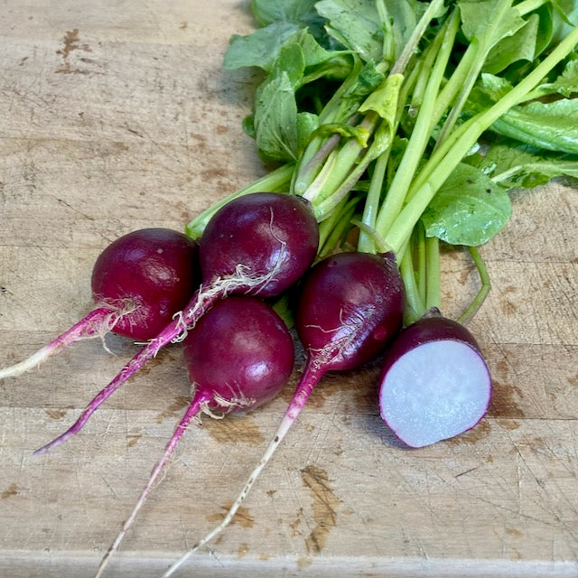 Cinq radis frais aux feuilles vertes sont posés sur une planche à découper en bois. Quatre d&#39;entre eux, probablement des Radis Malaga - semences bio de Tourne-Sol, ont la peau rouge et les racines attachées. L&#39;un d&#39;eux est coupé en deux, révélant son intérieur blanc.