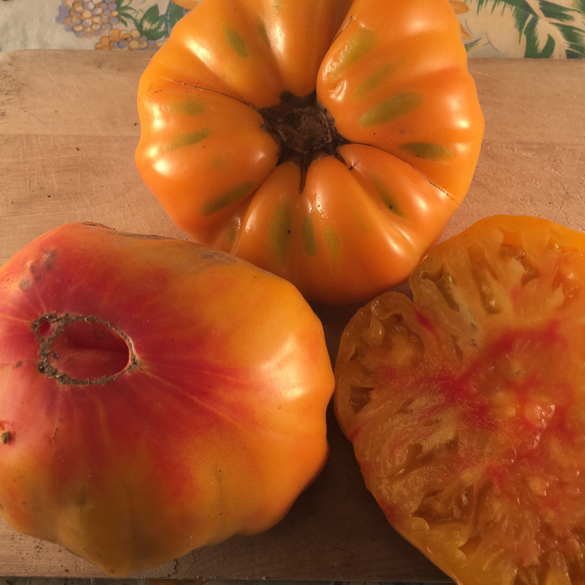 Trois tomates oranges allemandes mûres de Tourne-Sol reposent sur une surface en bois : deux sont entières, présentant des stries orange et rouges vibrantes, tandis qu&#39;une est tranchée pour révéler son intérieur juteux. Leur apparence brillante et leur goût fruité les rendent irrésistibles aux côtés de la tomate jaune subtilement sucrée.