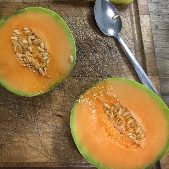 Un melon Oka - semences bio de Tourne-Sol coupé en tranches, ressemblant à un melon précoce, repose sur une planche de bois usée avec des graines visibles. Une cuillère en métal attend à proximité pour être récupérée. La planche et le plan de travail présentent des signes d&#39;utilisation régulière.